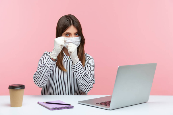 Fight coronavirus. Woman office employee sitting safe healthy with hygienic face mask and protective gloves during quarantine and keeping fists clenched to punch, boxing gesture. indoor, studio shot - Foto, afbeelding