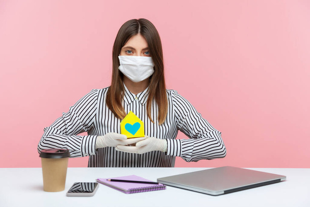 Renting during coronavirus quarantine. Woman real estate agency sitting at workplace, wearing hygienic mask and protective gloves, holding paper house, offering help in mortgage issues, home insurance - Foto, Bild