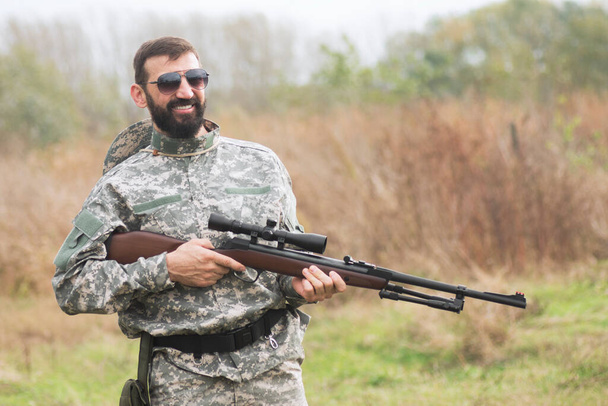 O soldado sorridente do exército de uniforme militar está segurando uma espingarda ou arma de fogo ao ar livre
. - Foto, Imagem
