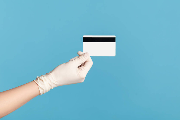 Perfil vista lateral close-up da mão humana em luvas cirúrgicas brancas segurando cartão de crédito clássico. indoor, studio shot, isolado em fundo azul
. - Foto, Imagem