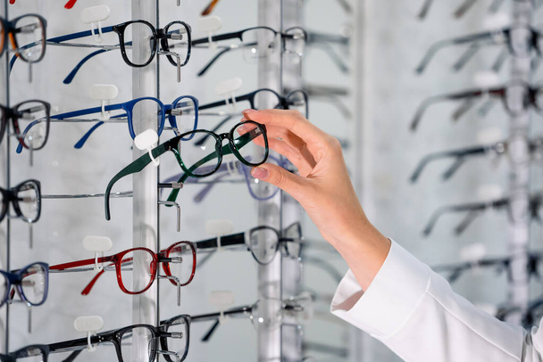 Row of glasses at an opticians. Eyeglasses shop. Stand with glasses in the store of optics. Woman's hand chooses glasses - Fotoğraf, Görsel