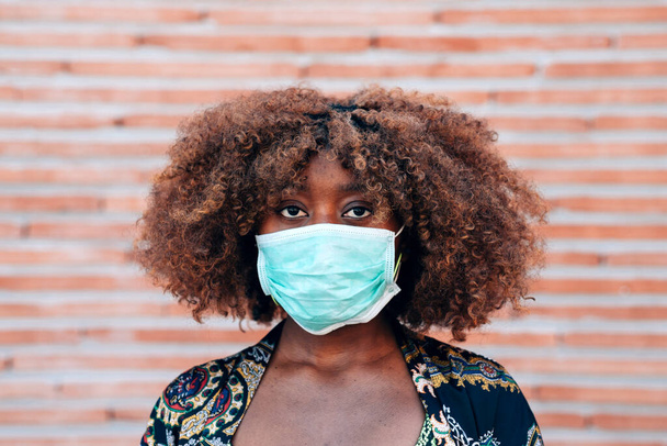 Close up portrait of young black woman wearing face medical mask to prevent coronavirus infection. bricks background. Protection against viruses - Foto, Imagem