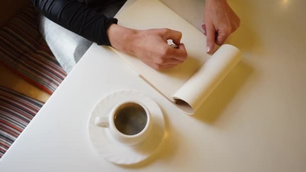 Bir kızın eli eskiz defterinde bir kafede kalemle, bir fincan kahveyi düz bir masada çizer. - Video, Çekim
