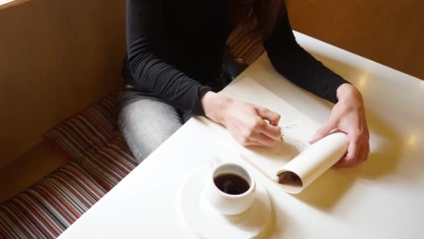Bir kızın eli eskiz defterinde bir kafede bir kalem, masanın üzerinde bir fincan kahve, sarmal bir şekilde en üstten kayan bir kamera çizer. - Video, Çekim