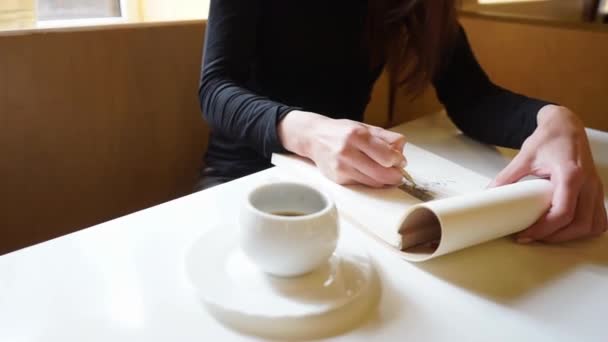 Bir kızın eli, eskiz defterinde bir kafede kalemle masanın üzerinde bir fincan kahve, yavaşça seyahat eden bir resim çizer.  - Video, Çekim