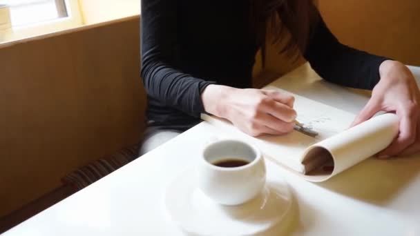 uma mão de uma menina desenha no caderno de esboços com um lápis em um café, uma xícara de café na mesa, tiro viajando lento
  - Filmagem, Vídeo