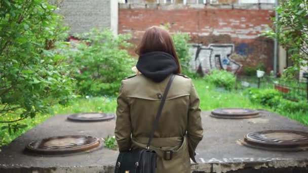 dziewczyna w płaszczu przeciwdeszczowym stoi tyłem do nas przed kolekcjonerem ścieków i murem z cegły - Materiał filmowy, wideo