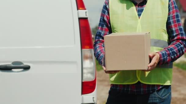 A courier with a cardboard box stands against the background of a white delivery service van - Materiał filmowy, wideo