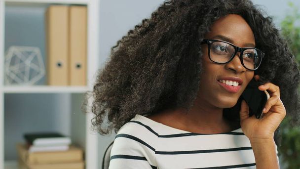 Primer plano de la joven mujer afroamericana bonita en gafas charlando en su teléfono inteligente en la oficina y sorprendiéndose. Retrato plano
. - Foto, Imagen