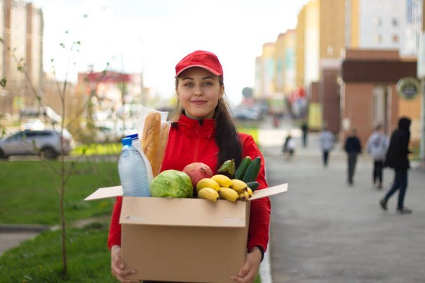 Ταχυδρόμος κορίτσι με ένα κουτί φαγητό στο δρόμο. Το κορίτσι από την υπηρεσία παράδοσης κατέχει ένα κουτί στο οποίο είναι: λαχανικά, φρούτα, ψωμί, νερό, γάλα. Κρατάει το κουτί μπροστά και χαμογελάει.. - Φωτογραφία, εικόνα