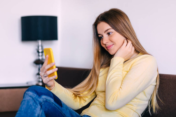 Beautiful young cheerful woman taking selfie with smile while sitting on couch - Фото, изображение