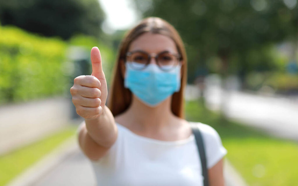 Joven estudiante con máscara quirúrgica que muestra un signo de como al aire libre. Concéntrate en la mano
. - Foto, imagen