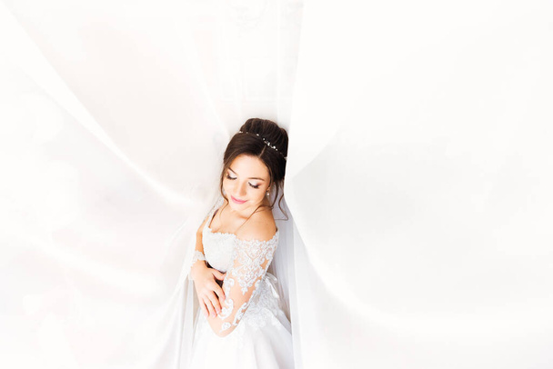 Mooie bruid in witte jurk sloot haar ogen op de achtergrond van witte gordijnen - Foto, afbeelding
