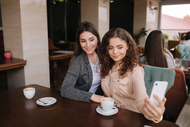 Atrakcyjne młode kobiety robiące selfie w restauracji. Kobiety drin cappuccino na tarasie. Dwie kobiety robią razem zdjęcia. - Zdjęcie, obraz