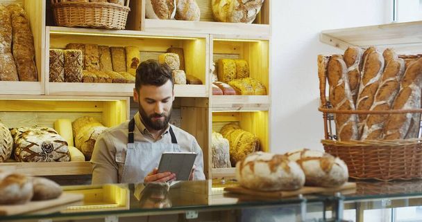 Portrait of male baker working in bakery shop and using tablet - 写真・画像