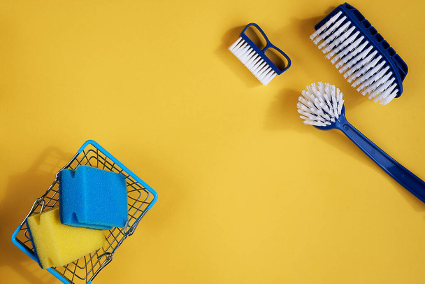 Blue dish brushes, sponges in metal basket, cleaning rag and yellow gloves and sponge on a yellow background. Space for text. top view - Photo, image