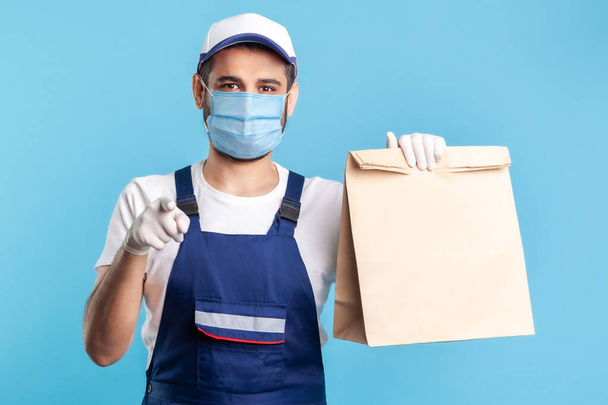 Hey you! Portrait of cheerful bearded handyman in overalls, mask and gloves carrying parcel, pointing to camera. Courier delivering food in paper bag, post mail services. indoor studio shot, isolated - Photo, Image