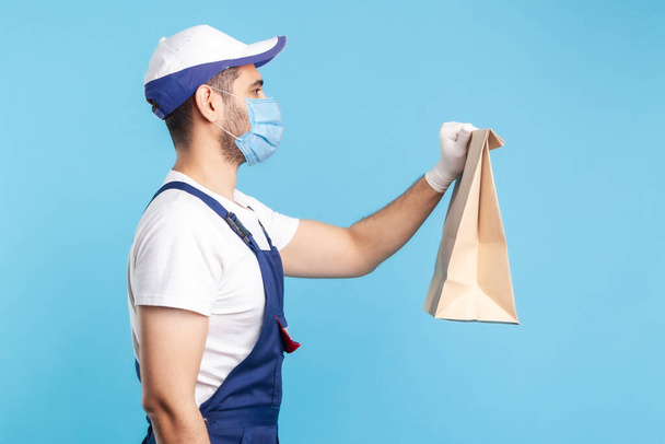 Side view, friendly professional courier in uniform, mask and safety gloves giving groceries bag, delivering takeaway food, post services express shipping. indoor shot isolated on blue background - Foto, immagini