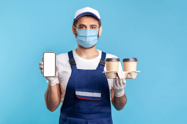 Delivery mobile app, internet advertise. Happy friendly courier man in overalls and mask holding coffee cups and cellphone with mock up display online order service. indoor isolated on blue background - Фото, изображение