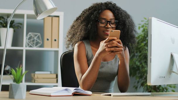 Гарна кучерява афроамериканська дівчина, що розмовляє і сміється на своєму смартфоні в сучасному офісі. Портрет - Фото, зображення