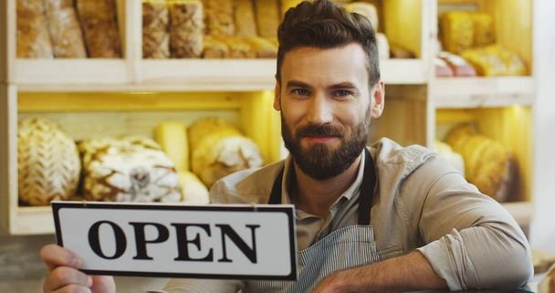 Portrait of handsome smiling baker holding an OPEN signboard in the bakery shop. - Photo, image
