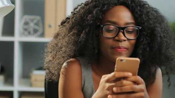 Close up of beautiful curly smiled African American young woman texting on her smart phone in the office. Portrait shot. Indoor - Felvétel, videó