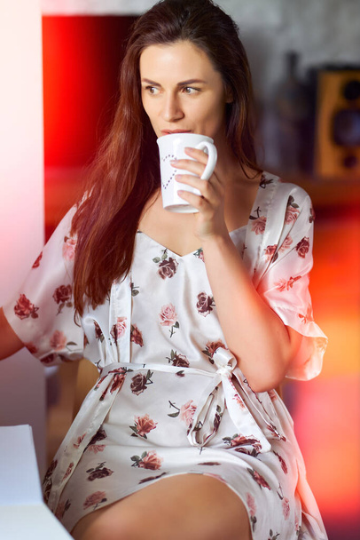 schöne Frau im Pyjama trinkt Wasser aus einer Tasse am Schreibtisch - Foto, Bild