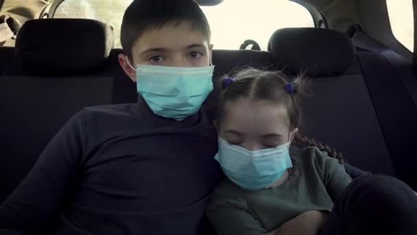 Κοντινό πλάνο παιδιά αγόρι και νυσταγμένο κορίτσι με ιατρική μάσκα κοιτάζει δυστυχώς στην κάμερα στο πίσω κάθισμα του αυτοκινήτου κατά τη διάρκεια της παγκόσμιας επιδημίας του ιού - Πλάνα, βίντεο