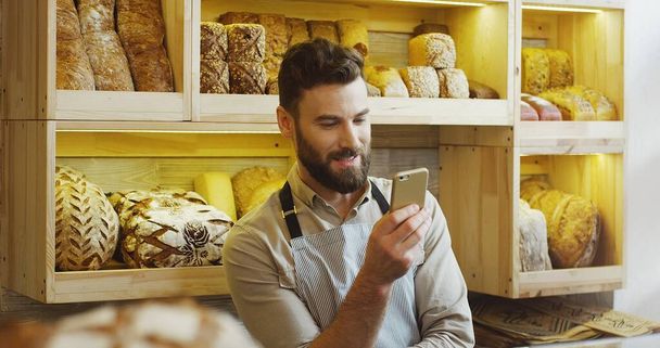 Portrait of happy baker working in bakery shop using smartphone. - Foto, imagen
