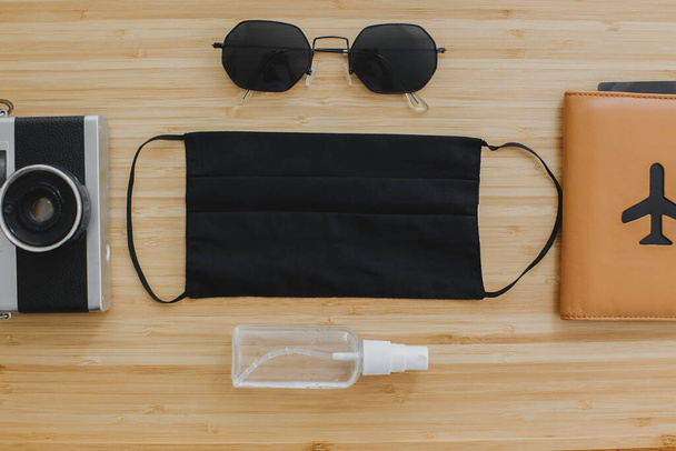 Viagens seguras no verão de 2020. Máscara preta, anti-séptico e desinfetante, passaporte, óculos de sol e câmera de foto no fundo de madeira flat lay. Medidas de proteção de férias Coronavirus
 - Foto, Imagem