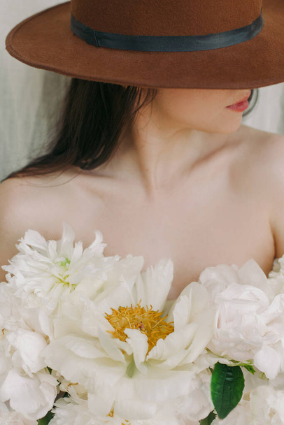 Virágos esztétika. Érzéki kép gyönyörű nő bazsarózsa csokorral ing helyett a bőrön. Illatot és öngondoskodást. Stílusos lány kalapban nagy fehér bazsarózsa virágok - Fotó, kép