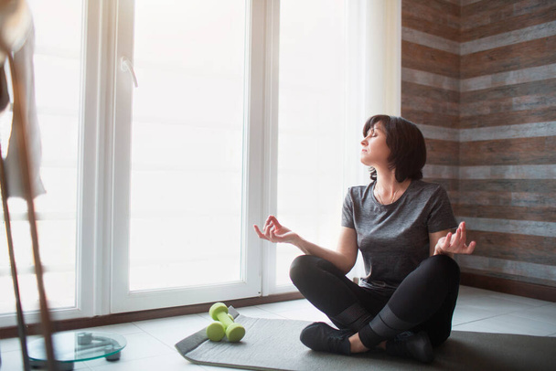 大人のフィットスリム女性は自宅でワークアウトを持っています。上級モデルの瞑想のプロセス。膝をついて窓を見て. - 写真・画像