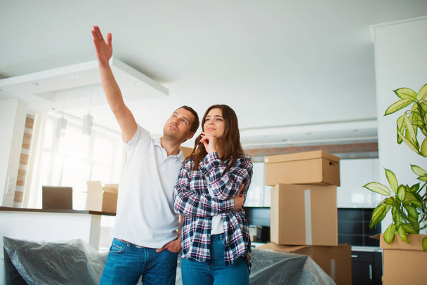 A young married couple in the living room in the house stand near unpacked boxes. hey are happy about new home. Moving, buying a house, apartment concept. - Photo, Image
