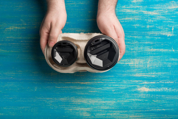 Dwie jednorazowe papierowe filiżanki kawy i uchwyt na kubek w męskich rękach na niebieskim drewnianym tle. - Zdjęcie, obraz