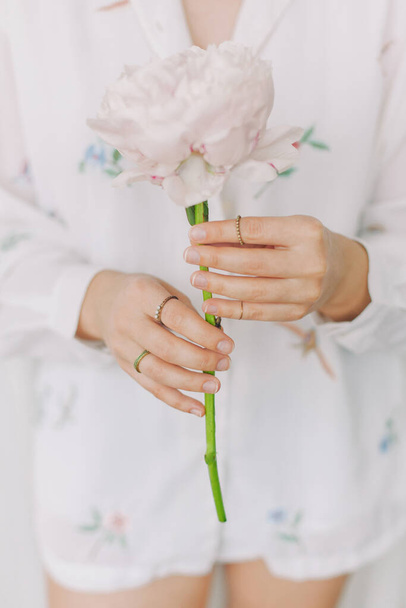 Κομψό κορίτσι σε λευκό floral πουκάμισο κρατώντας απαλά παιώνι λουλούδι στα χέρια με κοσμήματα. Αισθησιακή εικόνα. Όμορφη νεαρή γυναίκα κρατώντας μεγάλο ροζ παιώνια, περικοπή άποψη - Φωτογραφία, εικόνα