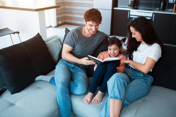 Aile evde kitap okuyor. Yetişkin erkek ve kız kardeş küçük kız kardeşe kitap okuyor. Genç anne-baba ve kız - Fotoğraf, Görsel