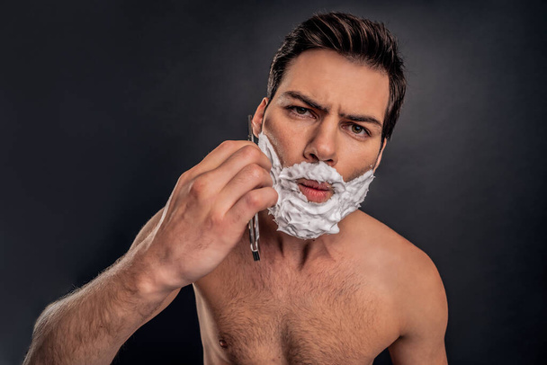 Młody człowiek z kremem do golenia na twarzy, pielęgnujący brodę brzytwą i patrzący w kamerę. - Zdjęcie, obraz