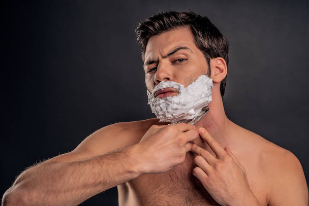 Mladý muž s krémem na holení na tváři, upravuje si vousy břitvou a dívá se do kamery. - Fotografie, Obrázek