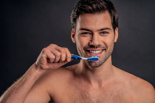 Handsome young bearded man isolated. Shirtless muscular man is standing on gray background with toothbrush and toothpaste in hand and smiling. Men care concept - Foto, afbeelding