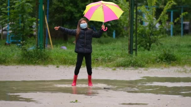Niña con un paraguas en una máscara médica y botas de goma salta a través de charcos en un día de primavera después de la lluvia. El concepto de protección de la infección por coronovirus covid-19
. - Metraje, vídeo