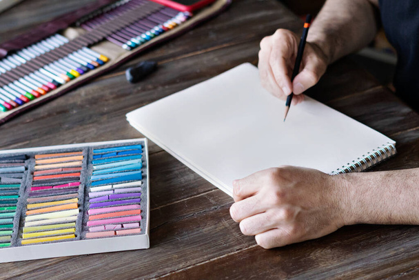 чоловік малює кольоровим олівцем і пастельними крейдяними чашами на папері. коробка з пастельних чаш і кольорових олівців на дерев'яному столі
 - Фото, зображення