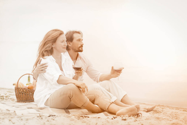 Charming couple drink wine and enjoy sunset on beach. Man and woman holds glasses of red wine sitting on the sandy beach while admiring beauty of sea or ocean. Romantic picnic concept. Toned image - Fotoğraf, Görsel