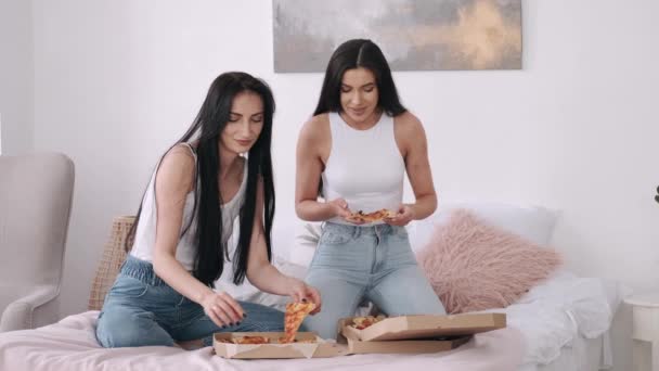 Two dark hair pretty ladies are sharing a takeaway pizza on the bed - Video