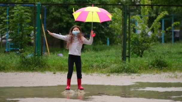 Mała dziewczynka z parasolem w masce medycznej i gumowymi butami skacze przez kałuże w wiosenny dzień po deszczu. Koncepcja ochrony przed zakażeniem covid-19 koronowirusem. - Materiał filmowy, wideo