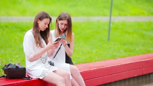 Twee jonge meisjes die buiten smartphones gebruiken. Twee vrouwen zitten in het park en sturen een boodschap per smartphone - Video