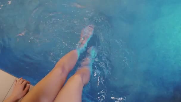 Close-up of female legs against the blue water by the pool. The girl dangles her legs in the water of the pool. Slow motion, 4K - Video