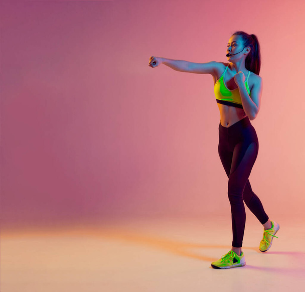 Cute girl fitness instructor teaches Boxing or body combat online training remotely on a bright neon background. - 写真・画像