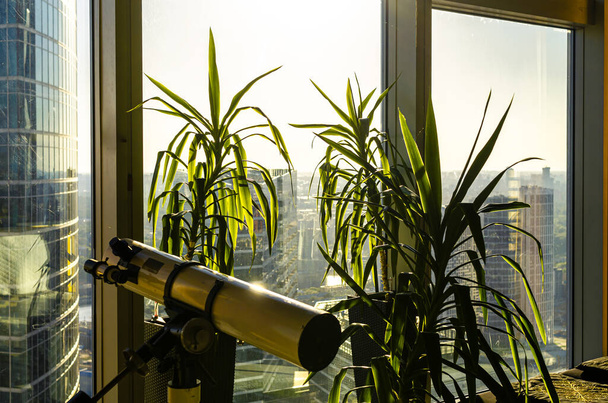 Панорамний вид через офісне вікно хмарочоса у бізнес-центрі з двома великими зеленими кімнатними рослинами та підсвічуванням телескопа з сонячним світлом на передньому плані
 - Фото, зображення