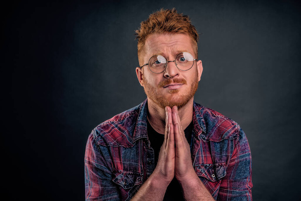 Close-up shot of redhead man in need standing with hands in pray over chest, pursing lips and frowning while asking forgiveness or begging for huge favour, being in troubled over gray background - Photo, Image