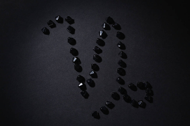Символ знака зодиака "Козерог", выполненный черными камнями на черном фоне. Низкий темный ключ. Зажигательное освещение. Тема гороскопа
 - Фото, изображение
